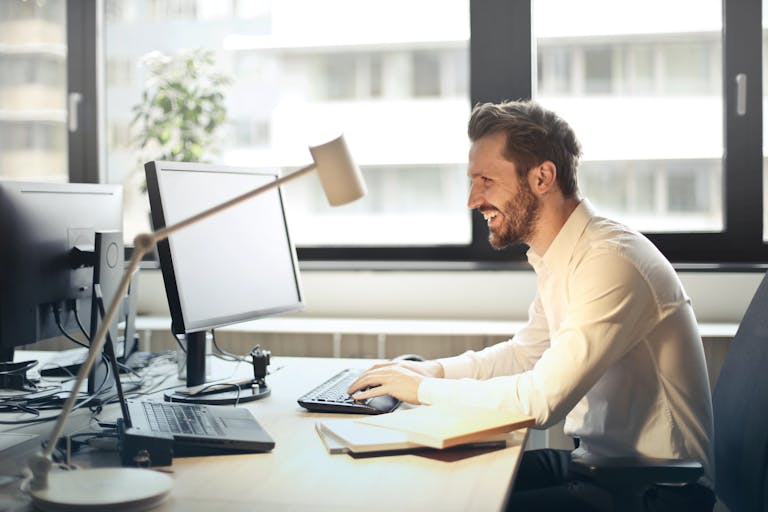 Ein Mann lächelt, während er an einem Büroschreibtisch mit einem Computer und natürlichem Tageslicht arbeitet, das durch große Fenster einfällt.